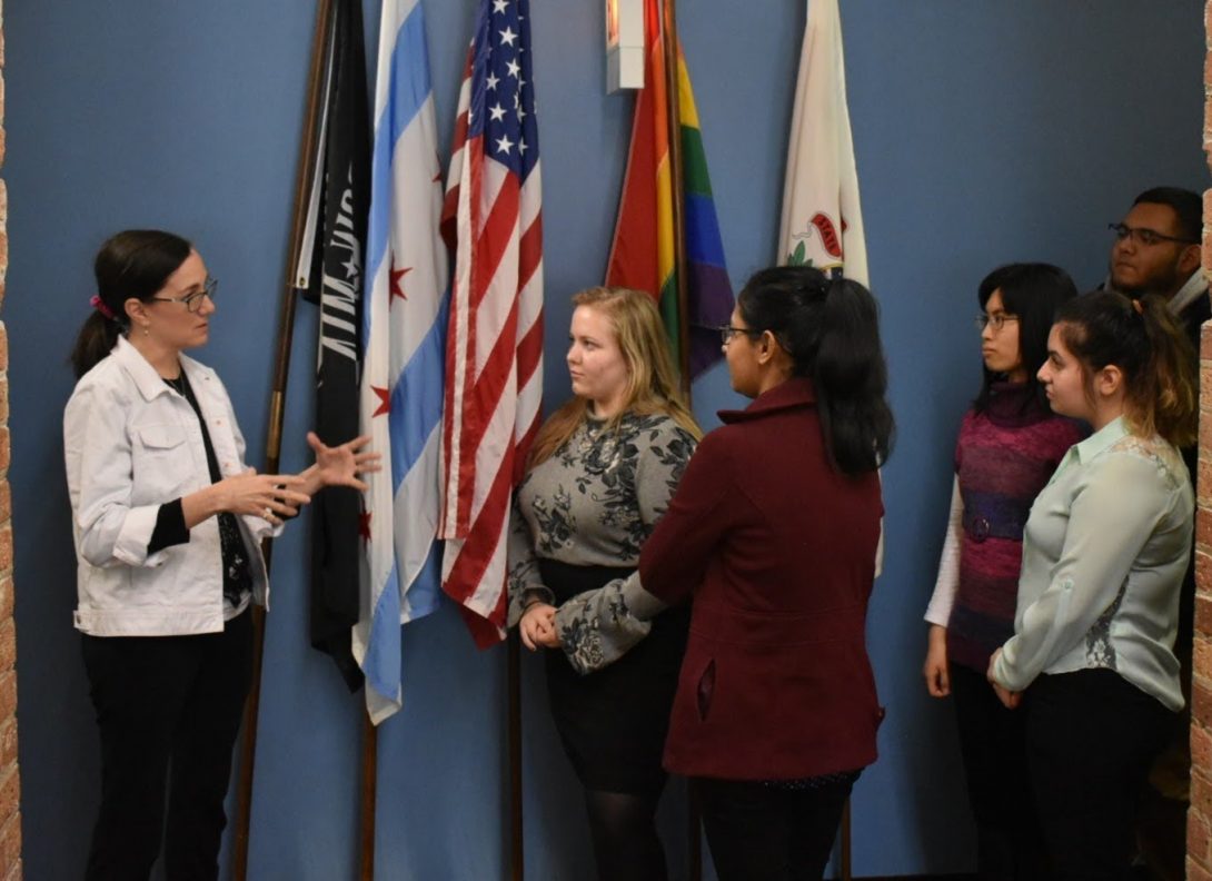 Representative discussing work environment to a group of students. In background, flags are displayed, including the City of Chicago flag and United States flag.
