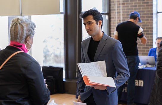 Student stands holding papers and speaking to tables at career event